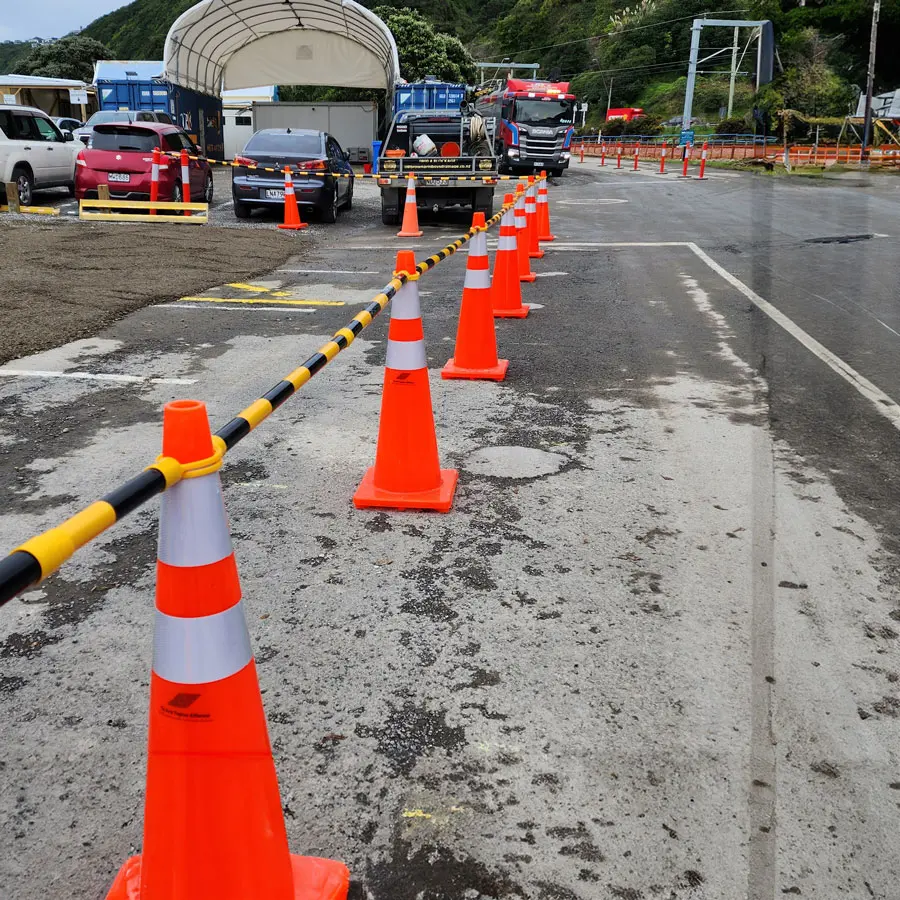 Line of orange traffic cones with reflective stripes, linked by yellow-black caution tape, guiding vehicles through a wet construction site with trucks and equipment in the background. Providing a traffic management solution.