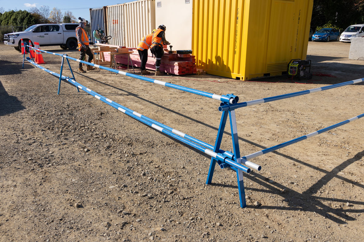 Long delineator pole run on A-Frame Stands, hi-vis workers behind.  Wide real-install of a long FRP delineator pole run supported on two blue Site Delineator A-Frame Stands across a gravel yard. Two workers in hi-vis at a materials stack near a yellow container behind. Stand variant ambiguous.