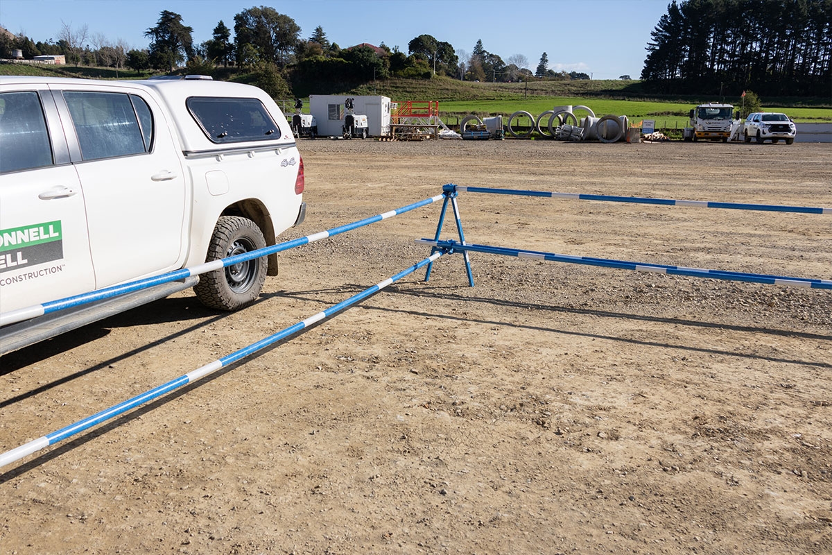 Site Delineator A-Frame Stand and poles cordoning a civil yard, ute at left.  Wide real-install on a gravel civil yard: a blue Site Delineator A-Frame Stand carrying long FRP delineator poles cordoning an area. White contractor ute at left with branding that appears to read McConnell Dowell; stacked concrete culverts and site huts behind. Stand variant ambiguous at distance.  u2014 Powerpac Image Intelligence v0.1 u00b7 candidates need review; possible new customer tag McConnell Dowell