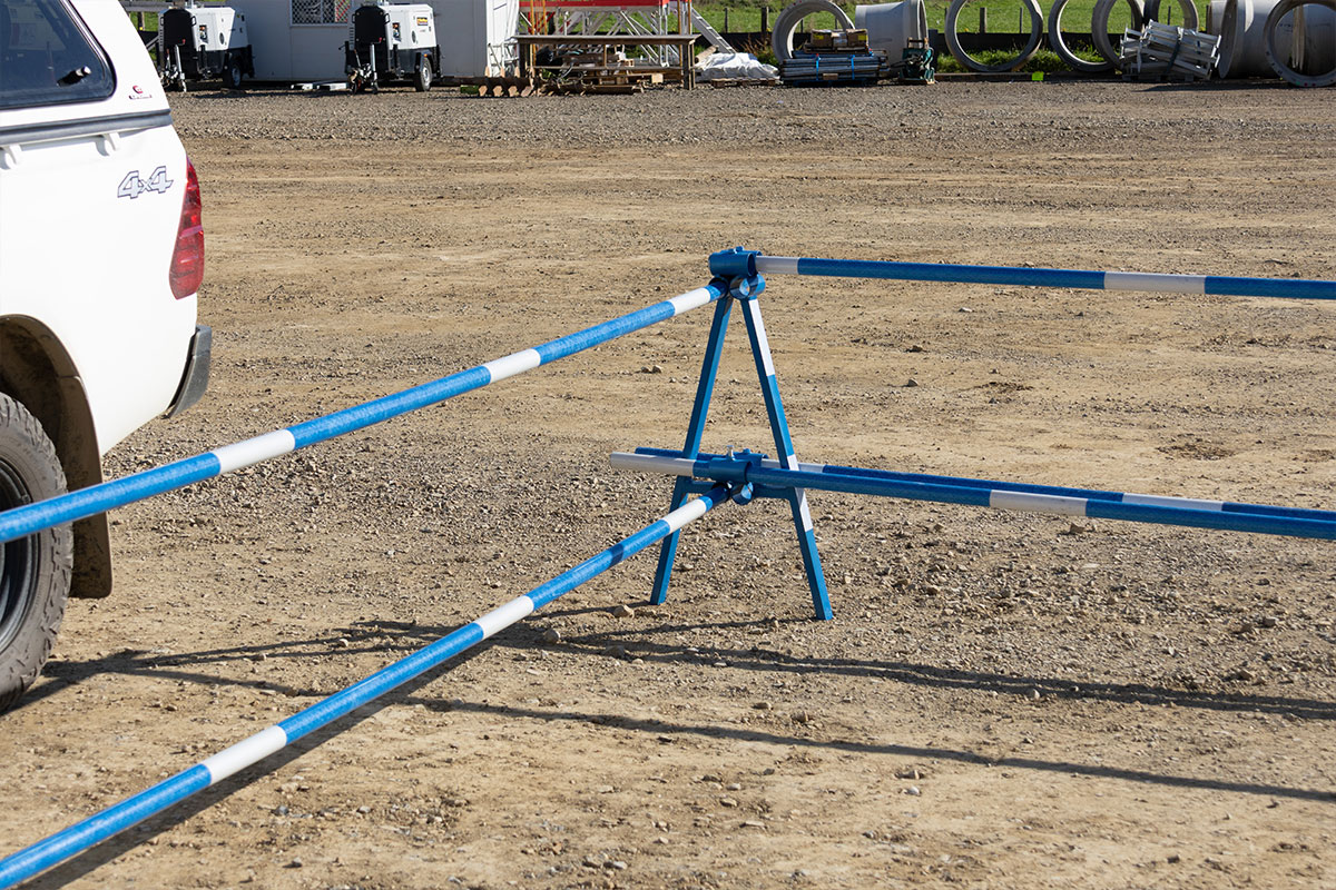 Delineator A-Frame Stand and pole run across a civil yard.  Wide real-install of a blue Site Delineator A-Frame Stand holding long FRP delineator poles across a gravel civil yard. White contractor ute at left